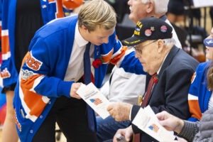 A high school student dressed in blue and orange is bent at the knees, facing an older man wearing a veteran's hat. The student has their hand on the veteran's shoulder, and is showing him a pamphlet with his other hand.