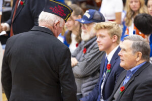 A standing veteran is speaking to a seated veteran. The standing veteran is wearing a garrison cap with red text that reads, "Past National Vice Commander 2023-2024," and the seated veteran has a red poppy pinned to their lapel.