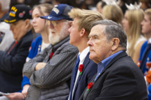 A line of veterans and student speakers sit, listening intently to the speech.