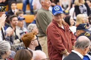 A veteran grandfather is standing and looking at his seated grandson as the crowd applauds the veteran for his service.