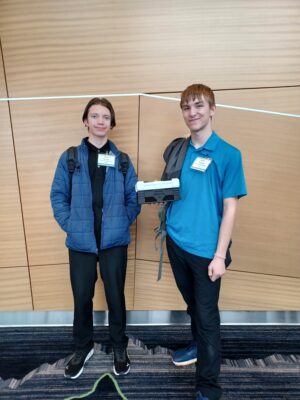 Two students wearing blue business casual outfits stand in front of a wooden wall backdrop. Both students are wearing backpacks, and one student is holding a plastic box.