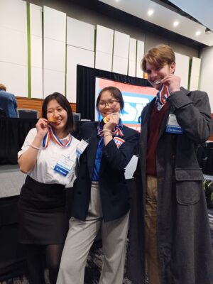 Three students wearing business casual stand in front of a large screen with the TSA logo. They are holding up respective gold medals.