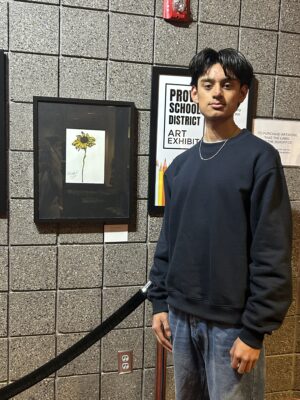 A male student stands next to his painting of a yellow flower.