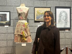 A female student stands next to a dress she has designed; the print is pink and black.