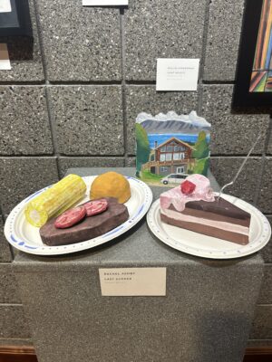 There is a clay and ceramic art piece of steak, corn, butter, and cake on two plates. Behind the ceramic art stands a fist-sized painting of a house.