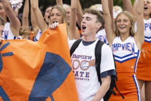 A boy holds a banner, cheering. Behind him, cheerleaders raise their hands.