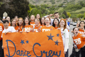 A group of students wearing orange and white shirts stand behind a large banner which reads, "Dance Crew."