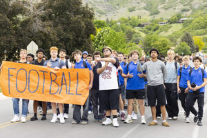 A group of students wearing orange and white shirts stand behind a large banner which reads, "Football."