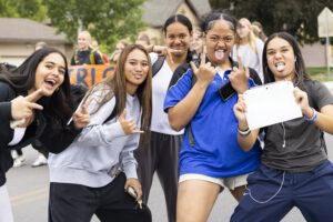 Five girls smile and stand together, throwing up peace signs and Shaka signs.