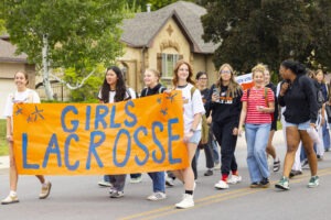 A group of students wearing orange and white shirts walk down a road, standing behind a large banner which reads, "Girls Lacrosse."