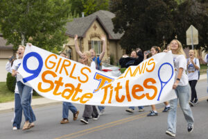 A group of female wearing white shirts stand behind a large banner which reads, "9 Girls Tennis State Titles."