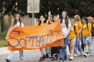 A group of students wearing orange and white shirts stand behind a large banner which reads, "Softball."
