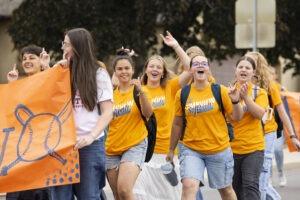 A group of students wearing orange shirts are smiling and waving at the camera.