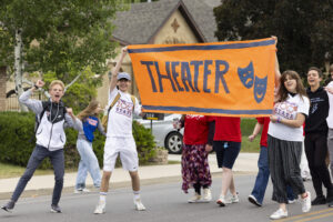 A group of students wearing orange and white shirts stand behind a large banner which reads, "theater." One student is jumping and cheering.