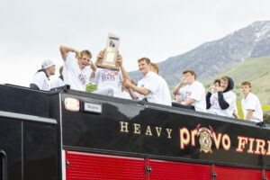 A group of students in white t-shirts are sitting atop a fire truck. They are holding up a state championship trophy.